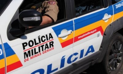 Viatura da Polícia Militar de Minas Gerais com um policial fardado ao volante, vista parcial do veículo com logotipo e cores institucionais visíveis.