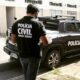 Agente da Polícia Civil de Minas Gerais em pé próximo a uma viatura oficial durante operação em área urbana, com prédios ao fundo.